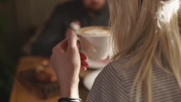 Молодая женщина держит чашку горячего капучино в руке, она ест в ресторане — стоковое видео
