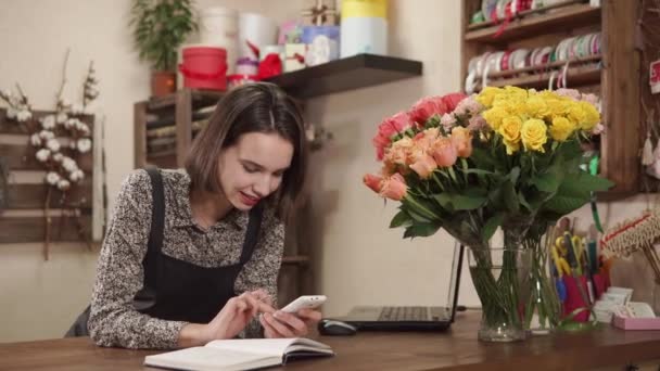 Ung kvinna arbetar som florist i en blomsteraffär och hon accepterar beställningar på buketter — Stockvideo
