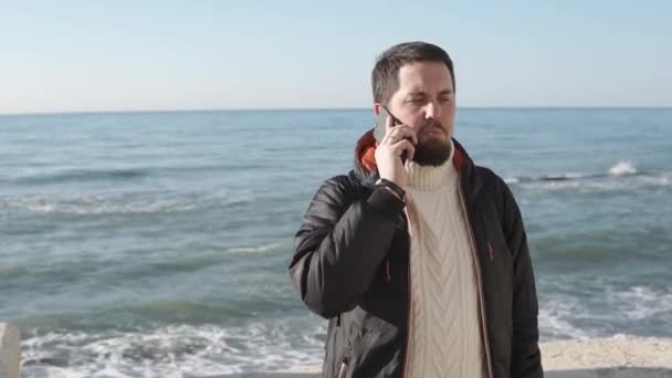 Дорослий чоловік, одягнений в осінню куртку, розмовляє на мобільному телефоні біля моря — стокове відео