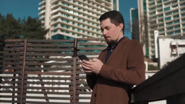 Hombre guapo está comprobando el correo electrónico por teléfono inteligente en una calle cerca de edificios altos — Vídeo de stock