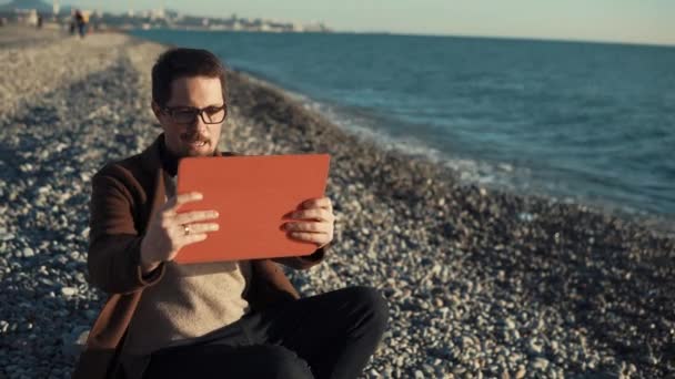 Sonriente hombre está haciendo videollamada por tableta en una playa de guijarros, agitando la mano — Vídeos de Stock