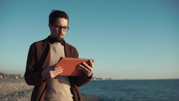 Barbudo hombre está utilizando el ordenador de cama plana sentado en la orilla del mar con panorama de la ciudad — Vídeos de Stock