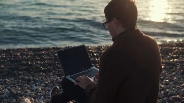 Un hombre adulto se sienta en una playa de piedra cerca del mar y recoge el texto en un ordenador portátil — Vídeos de Stock
