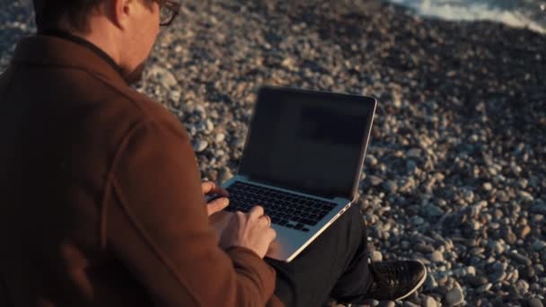 L'uomo sta lavorando con un computer portatile sedersi sulla riva di ghiaia del mare in serata — Video Stock