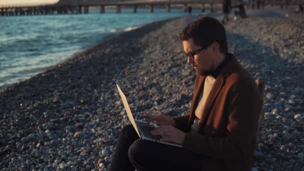 Felnőtt ember ül a köves strand, a naplemente alatt ember részt programozás — Stock videók