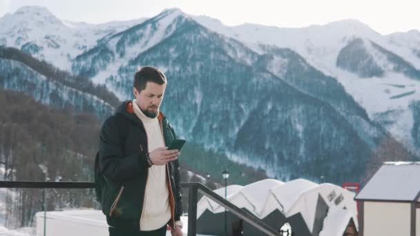 Человек, использующий мобильный телефон для отправки сообщений, стоящий на улице на горнолыжной базе в горах — стоковое видео
