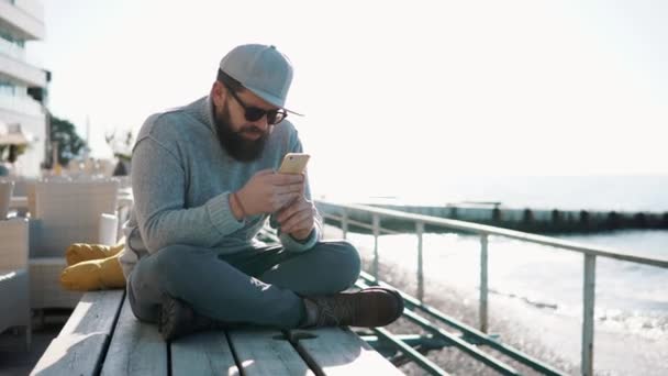 Junger Mann sitzt auf Holzbrettern am Meer und nutzt das Internet auf seinem Handy — Stockvideo