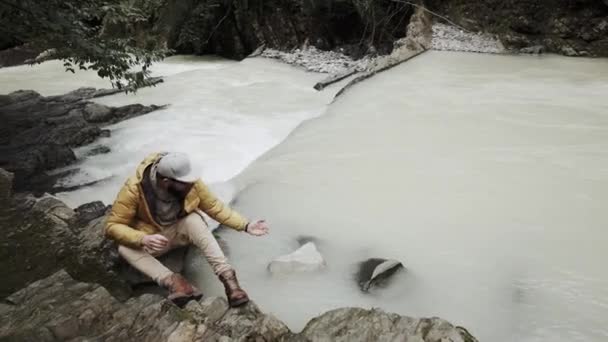 Un joven con barba se sienta a los pies de la cascada y se hace el tonto. — Vídeo de stock