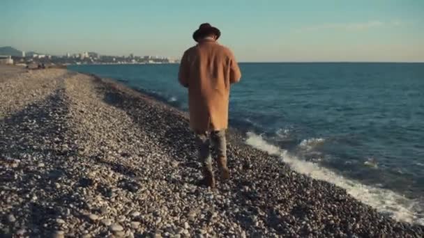 Un uomo con un lungo cappotto e cappello cammina lungo la riva, le onde battono — Video Stock