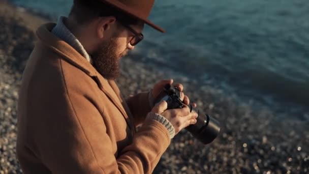 Een volwassen man zit op de kust en kijkt naar de monitor van zijn camera — Stockvideo