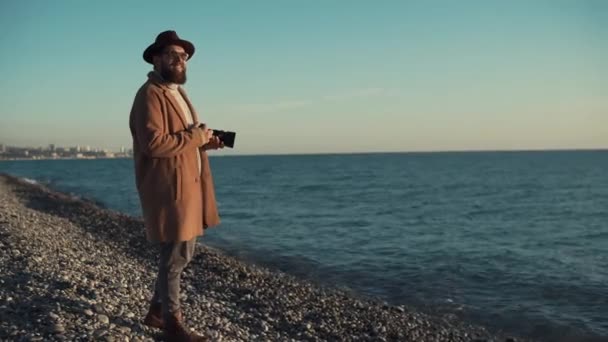 Νεαρό και κομψό φωτογράφος είναι κοντά στη θάλασσα και φαντάζεται πώς να φωτογραφίσει — Αρχείο Βίντεο