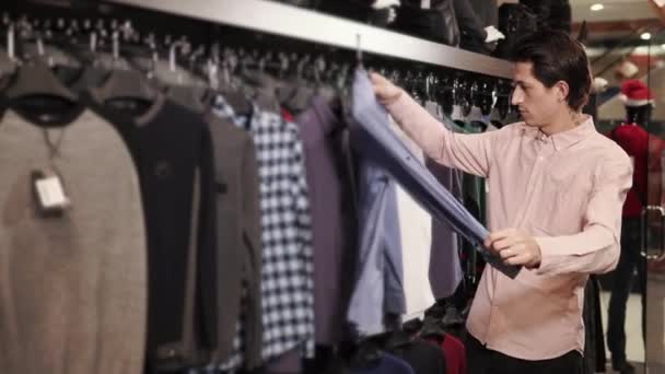 Um jovem olha para roupas em cabides, ele escolhe uma camisa em uma loja de roupas — Vídeo de Stock