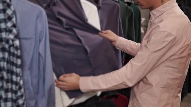 De mens is in een kleding winkel bestudeert kledingstuk op hanger, vinden van shirts — Stockvideo
