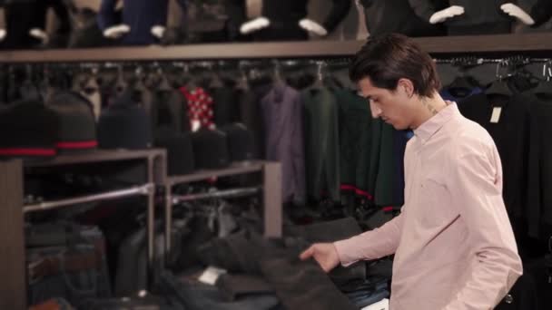 Молодой человек выбирает джинсы для ног, он в большом торговом центре — стоковое видео