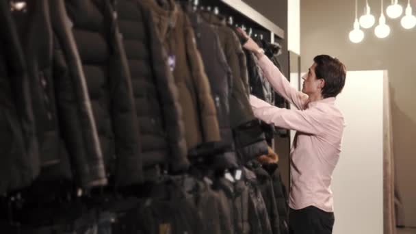 Чоловік бере чорну куртку з вішалки в магазині і носить на собі — стокове відео