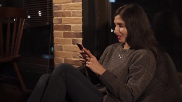晚上, 黑发女郎坐在咖啡豆袋椅上, 使用智能手机 — 图库视频影像