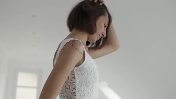Романтична брюнетка дівчина стоїть збоку і торкається її волосся в білій кімнаті — стокове відео