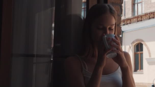 Nő iszik kávét ül az ablakpárkányon, és nézi az utcán — Stock videók