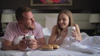 man and woman are eating croissants with coffee lying in bed in morning