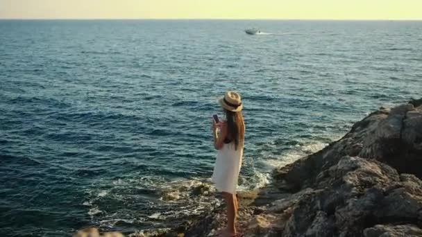 Mujer está tomando fotos de agua de mar por teléfono inteligente de pie en la orilla de la roca — Vídeos de Stock