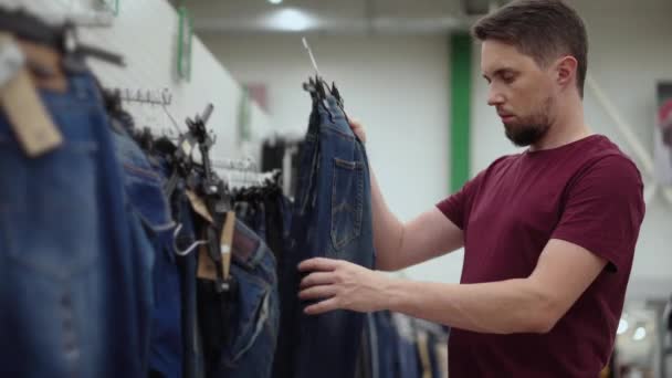 Hombre está eligiendo jeans en tienda de ropa, aplicando a su cuerpo — Vídeos de Stock