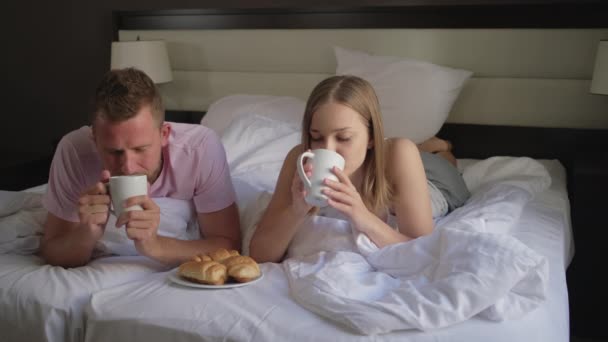 Mann und Frau liegen im Bett und trinken morgens Kaffee — Stockvideo