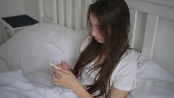 Νεαρή γυναίκα που μένει στο κρεβάτι στο ρεπό της και χρησιμοποιεί κινητό — Αρχείο Βίντεο