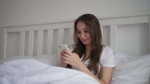 Mujer está leyendo mensajes en el teléfono móvil acostado en la cama por la mañana — Vídeos de Stock