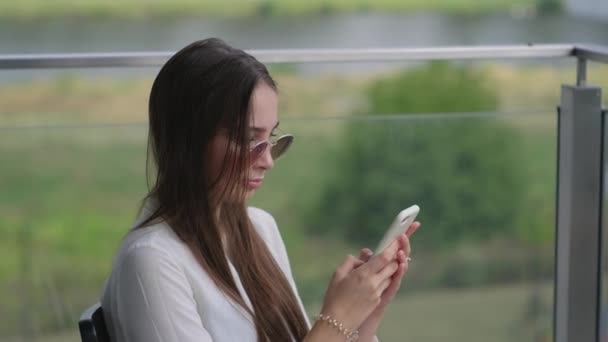Mujer joven está escribiendo sms en el teléfono inteligente sentado en la terraza en verano — Vídeos de Stock