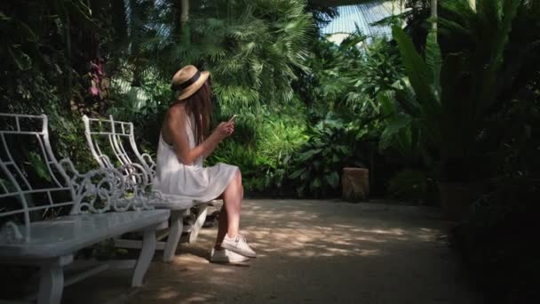 Жінка друкує повідомлення в смартфоні, сидячи всередині ботанічного саду — стокове відео
