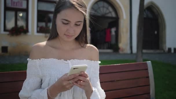 Mujer bonita está escribiendo sms en el teléfono celular sentado al aire libre — Vídeos de Stock