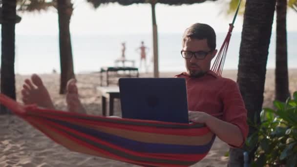 Мужчина использует ноутбук, лежащий в гамаке между ладонями — стоковое видео