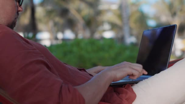 Человек расслабляется в гамаке, но делает работу на ноутбуке — стоковое видео