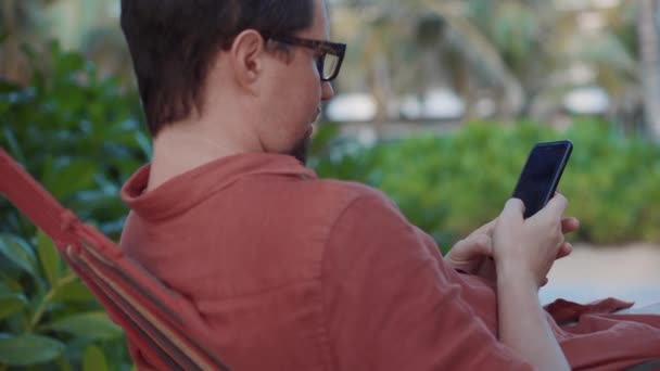Mobiles Internetsurfen. Mann mit Zelle entspannt sich in Hängematte — Stockvideo
