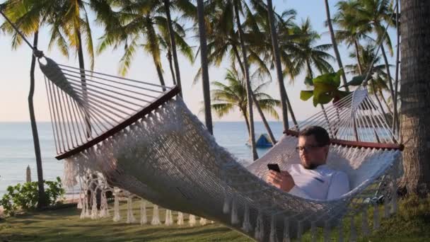 Muž s chytrým telefonem je relaxační v houpací síti — Stock video
