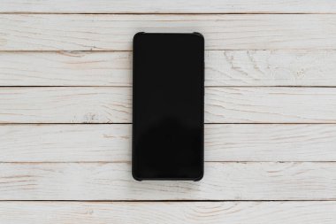 Beyaz ahşap arkaplanının ortasında koyu ekran olan akıllı telefon