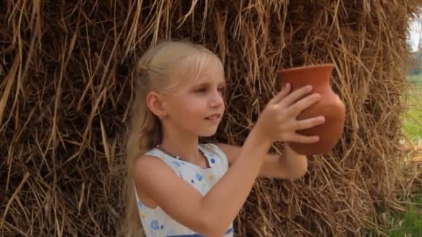 Leuk blond meisje drinkt verse koemelk uit een klei kruik tegen hooiberg in de zomer op de boerderij. Moeder geeft melk van een lachende meisje in een dorp. — Stockvideo