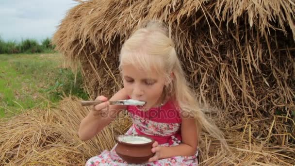 Χαριτωμένο ξανθό κορίτσι τρώει γιαούρτι γλυκό από ένα πήλινο καπάκι. — Αρχείο Βίντεο