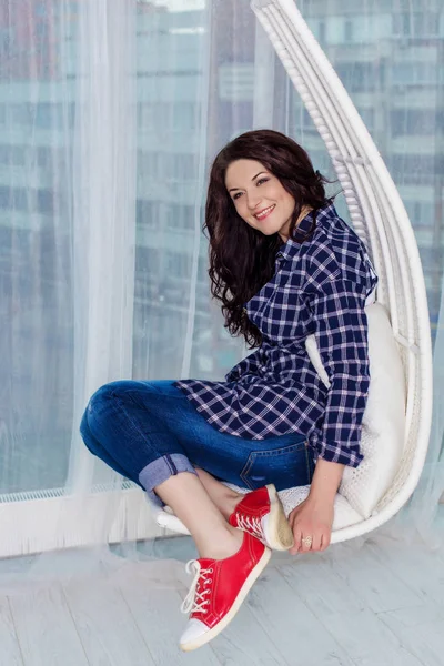 Yağma tarzı. Hippi kız kameraya gülümseyen ve salıncakta oturuyor. Siyah kıvırcık saçlı mavi kareli gömlek, kot pantolon ve kırmızı spor ayakkabı giyen genç beyaz kız.