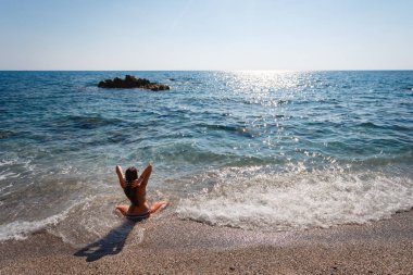 Denize karşı sahilde kum mavi mayo oturur uzun saçlı bir kız arkası seksi, küçük, zevk, turizm çakıl taşı. Rahatlatıcı ve tropikal plaj destinati zevk mutlu kaygısız kadın