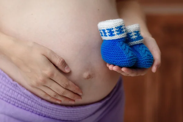 孕妇抱着蓝色婴儿靴子在肚子，特写 — 图库照片