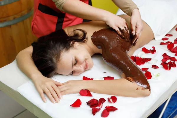 Masajista aplicando chocolate a la espalda de la mujer. Masaje en el salón de spa — Foto de Stock