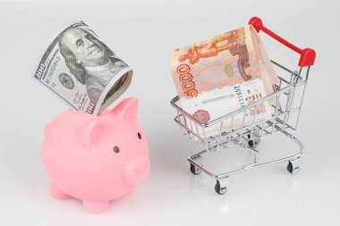 Pink piggy money box, dollar and rouble banknotes in mini shopping cart on gray background