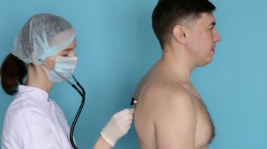 Maskeli kadın doktor fonendoskoplu bir adamın nefesini dinliyor.