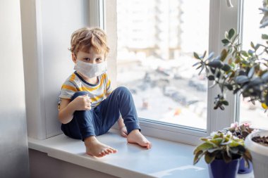 Maskeli çocuk pencerenin yanında oturuyor ve bakıyor. Evde kalın, Coronavirus Covid-19 karantinası