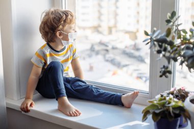 Maskeli çocuk pencerenin yanında oturuyor ve bakıyor. Evde kalın, Coronavirus Covid-19 karantinası