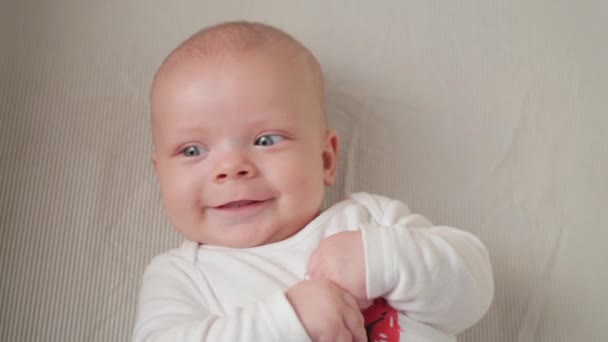 A cute little baby is looking into the camera and is happy on a white bed sheet. — Stock Video