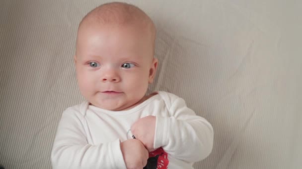 Um bebê pequeno bonito está olhando para a câmera e está feliz em um lençol branco . — Vídeo de Stock