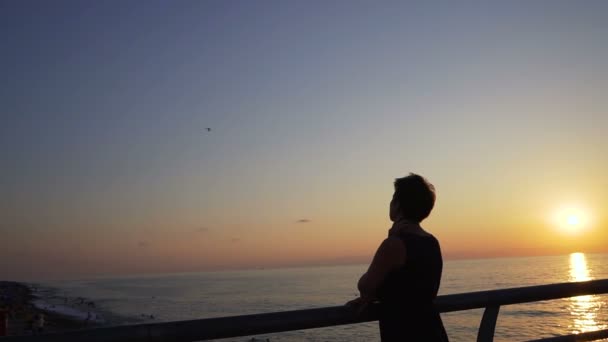 Jovem mulher fica na orla e olha para o pôr do sol — Vídeo de Stock