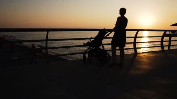 Мать с коляской идет вдоль вечерней набережной у моря — стоковое видео